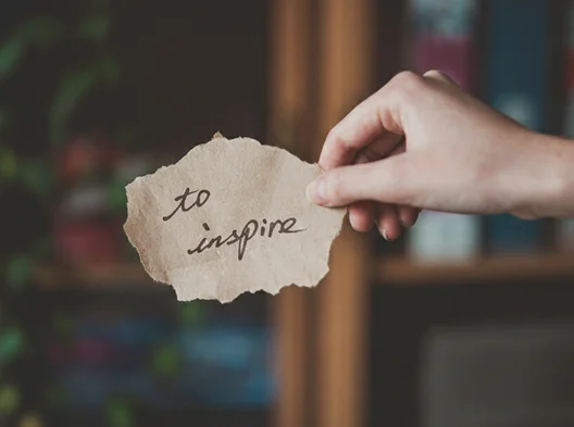 Hand holding a note with the words “To Inspire,” symbolizing creativity and storytelling.